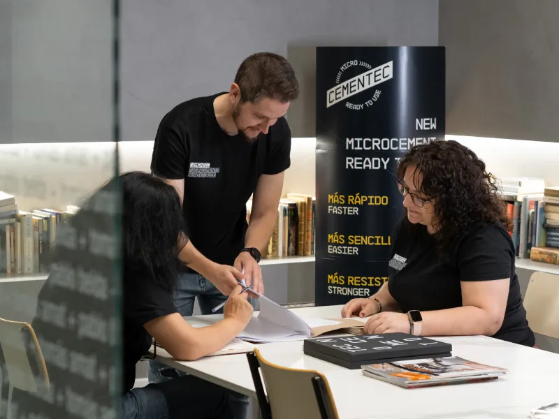 Three professionals in black Cementec t-shirts collaborate over documents at a white table in a modern office. A man stands and points to a folder while a woman signs a paper, with a promotional banner for Cementec microcement and bookshelves in the background.